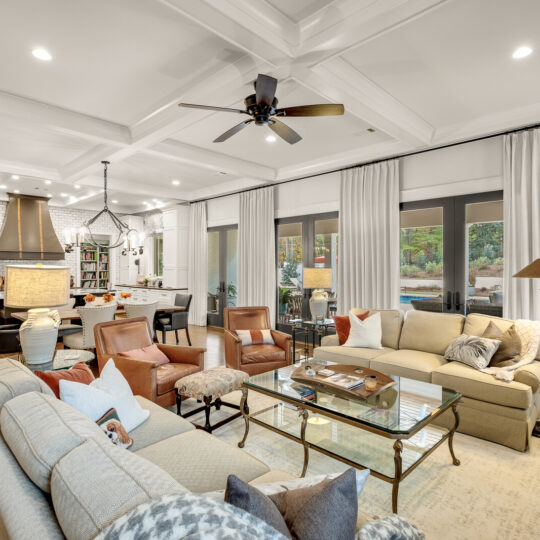 Spacious living room with modern decor featuring light-colored sofas, armchairs, and a glass coffee table. An open kitchen with a large island is visible in the background. Large windows overlook a patio area, and the room is lit by recessed lighting and a ceiling fan.