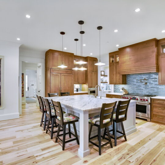 Spacious kitchen with wooden cabinets, marble island, and tiled backsplash. Five chairs surround the island. Pendant lights hang above. Framed artwork decorates the wall. Warm, natural wood flooring throughout.