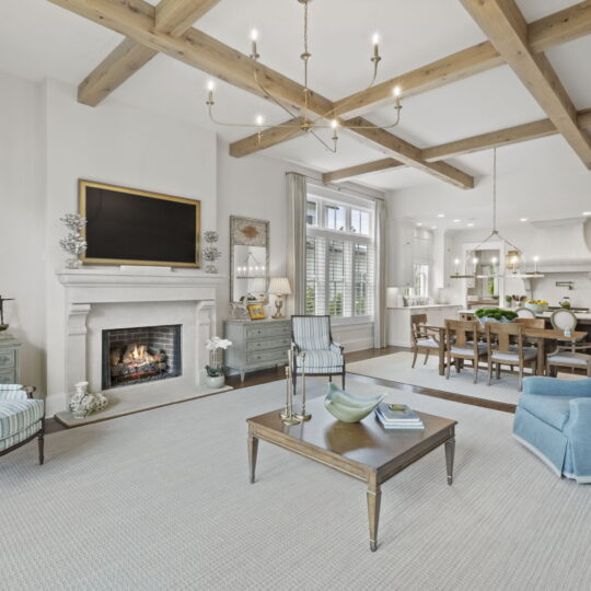 Spacious living room with white walls, wooden beam ceiling, and a lit fireplace. Includes blue and striped chairs, a wooden coffee table, and a large television above the fireplace. Dining area and kitchen with modern decor visible in the background.