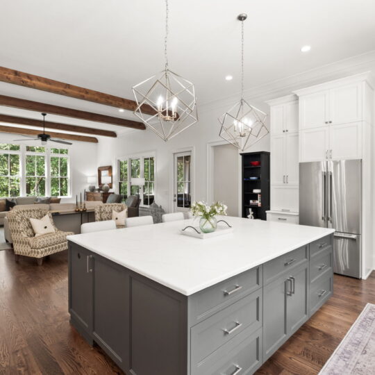 Spacious open-concept kitchen and living room featuring a large gray island with a white countertop. Modern chandeliers hang above, and exposed wooden beams add character. A pantry with organized shelves is visible on the right, and large windows illuminate the space.