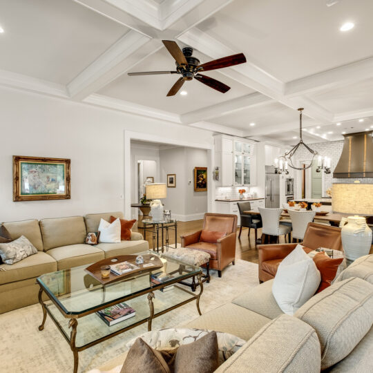 Spacious living room with beige sofas, a glass coffee table, and decorative pillows. A ceiling fan is above. The kitchen and dining area are visible with a chandelier and modern appliances. Walls are adorned with artwork. Cozy and well-lit space.