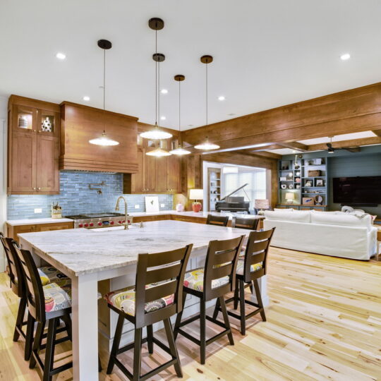 Spacious kitchen and living area with wooden floors and ceiling beams. A large marble island with barstools centers the kitchen, surrounded by wooden cabinets. Cozy living room with sofas and a fireplace is visible in the background.