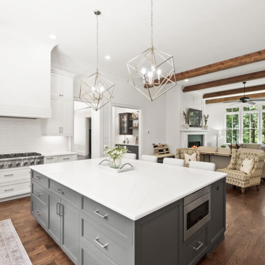 A modern kitchen with a large gray island featuring white marble countertops. Two geometric pendant lights hang above. The background shows a living area with a sofa, chair, and a fireplace. Wooden beams accentuate the white ceiling.