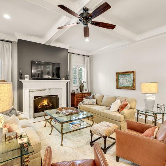 A cozy living room with a fireplace, beige sofas, and armchairs. A TV is mounted above the fireplace. The room features a ceiling fan, two glass coffee tables, decorative lamps, and a painting on the wall. A hallway is visible on the right.