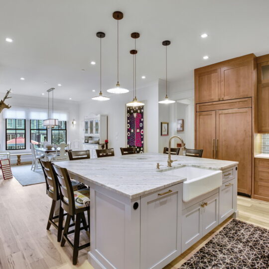 Spacious kitchen with a large island featuring a white countertop and farmhouse sink. Pendant lights hang above. Theres a brick accent wall with artwork, wooden cabinets, and a dining area in the background. Light wood flooring runs throughout.