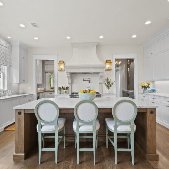 Spacious kitchen with white cabinets, a large island with a wooden base, and four cushioned chairs. Elegant range hood, recessed lighting, and hardwood floors add warmth. A bowl of fruit and flowers adorn the counters, and windows bring in natural light.