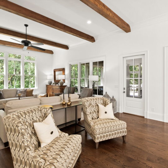 A spacious living room with wooden floors, patterned armchairs, and a beige sofa set. Wooden ceiling beams add warmth. Large windows bring natural light, offering a view of lush greenery outside. A ceiling fan and framed artwork complete the decor.