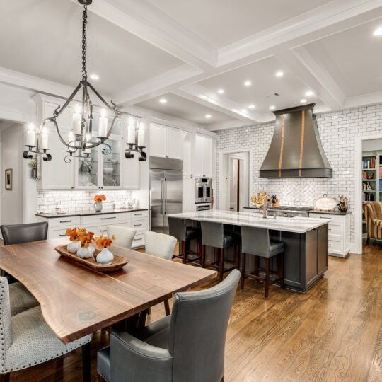 Elegant kitchen with white cabinets, brick backsplash, and stainless steel appliances. A large island features barstools, while a wooden dining table with upholstered chairs sits beneath a chandelier. A cozy room with bookshelves is visible in the background.