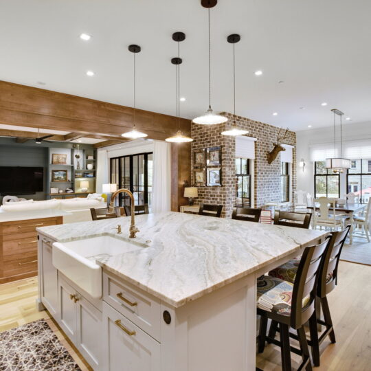 A spacious kitchen features a large marble island with a farmhouse sink. Pendant lights hang above the island. Wooden cabinets and brick accents decorate the space. A dining area with windows is visible in the background, offering ample natural light.