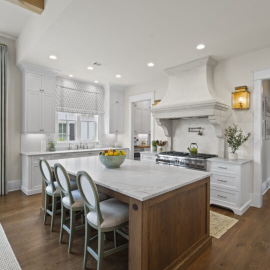 A spacious, modern kitchen with white cabinetry, a large marble island, and four chairs. A bowl of fruit sits on the island. Stainless steel appliances and a white range hood are visible. Sunlight streams through large windows with light curtains.