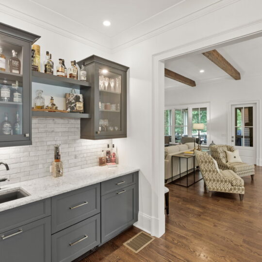 A modern home bar area with gray cabinets, a marble countertop, and a small sink. Bottles of liquor are displayed on the counter and shelves. In the background, theres a cozy living room with armchairs and a window view.