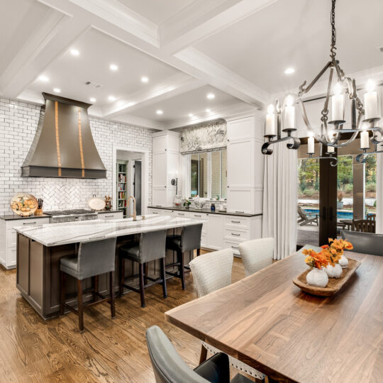 A modern kitchen features a large island with gray chairs, a stainless steel hood, and white subway tile backsplash. A dining table with a chandelier hangs nearby. The room has hardwood floors, white cabinetry, and large windows.