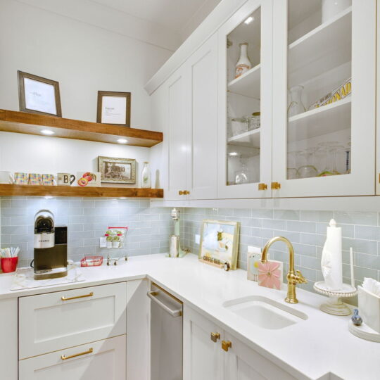 A modern kitchen with white cabinets and a light blue tiled backsplash. It features a sink with a gold faucet, a coffee machine, and glassware on display. Wooden shelves hold decorative items, and a patterned rug covers the floor.