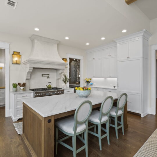 A spacious kitchen with a large island featuring four cushioned chairs. The island has a marble countertop. The room has white cabinets, a prominent range hood, and wood flooring. A doorway leads to an adjoining room with more cabinetry.