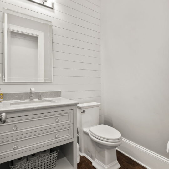 A modern bathroom featuring a gray vanity with a marble countertop, a rectangular mirror, and overhead lighting. A white toilet is beside the vanity. The walls are white shiplap, and the floor is dark wood. A woven basket is on a lower shelf.