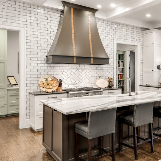 A modern kitchen with a large island featuring a marble countertop and four dark upholstered stools. The walls are lined with white subway tiles, and a prominent black range hood is above the stove. Theres ample cabinetry and a view into an adjoining room.