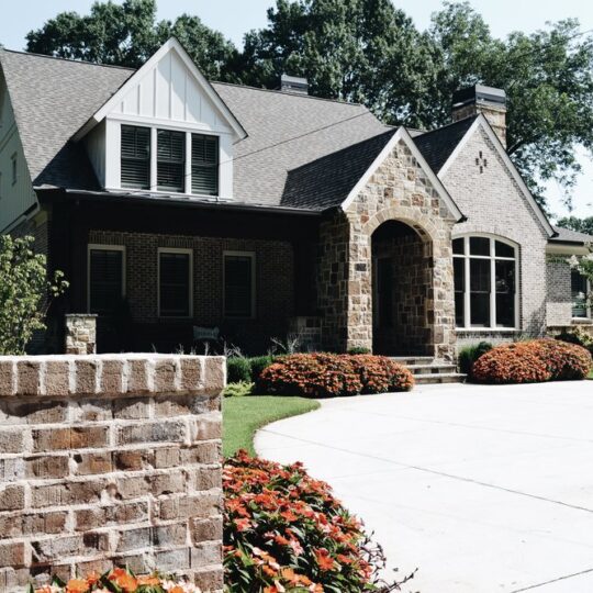 A quaint house with a gabled roof and stone facade, surrounded by lush greenery and vibrant flower beds. The driveway is wide and paved, leading up to the entrance. Tall trees are visible in the background.