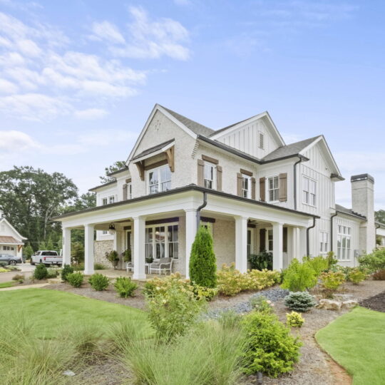 A large white two-story house with a wraparound porch and gabled roof. The well-maintained lawn features bushes, small trees, and a flower bed. The sky is partly cloudy, and there is another house visible in the background.