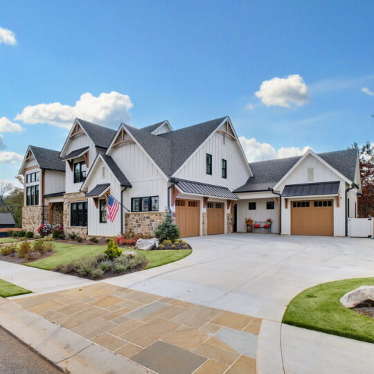 Modern suburban house with a stone and white exterior, multiple gables, and a three-car garage. An American flag hangs near the entrance. The driveway is expansive, and the front yard is landscaped with shrubs and young trees. Sky is partly cloudy.