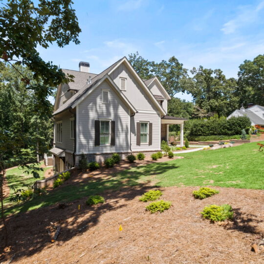 A light gray, two-story house with a gabled roof is nestled among trees and shrubs. Its situated on a sloped lawn with well-maintained landscaping. A neighboring house is visible in the background under a clear blue sky.