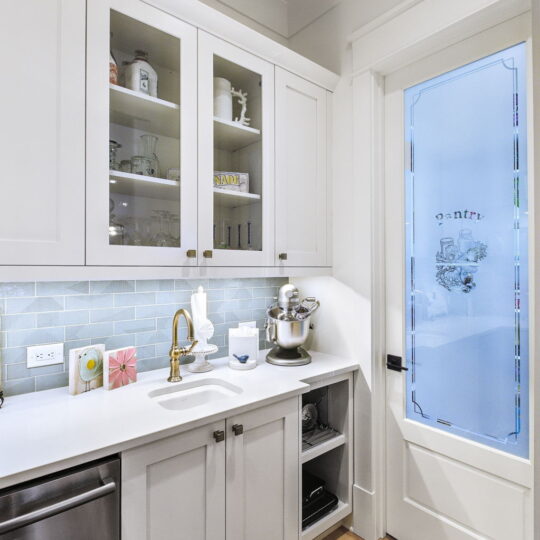 A modern kitchen with white cabinets and glass doors displaying dishes. A counter with a mixer, gold faucet, and sink. A frosted door labeled pantry leads to another room. The floor is wooden, and a patterned rug adds contrast.