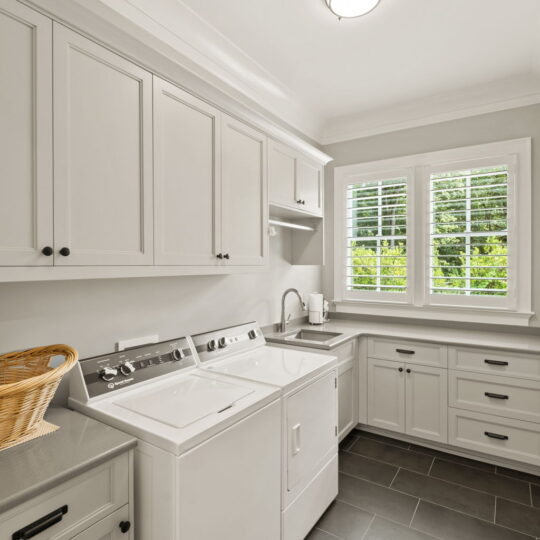 A bright laundry room with light gray cabinets and a dark tiled floor. There is a washing machine and dryer below the countertops. A woven basket is on top of a cabinet. A window with white shutters lets in natural light.