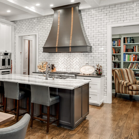 A modern kitchen with white cabinets, a dark island with four black stools, and a stainless steel refrigerator. The backsplash is white subway tile. A large black range hood is above the stove, and a cozy reading nook with books is visible through an open doorway.