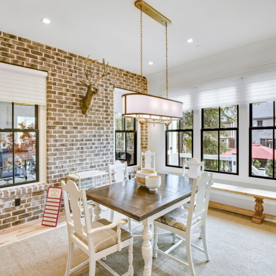 A cozy dining room with a brick accent wall features a wooden table and white chairs. A modern rectangular light fixture hangs above. Large windows let in natural light, and a deer head decorates the wall. A bench sits by the windows.