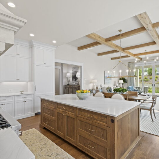 Spacious kitchen and living area featuring a large wooden island, white cabinetry, and exposed wooden beams on the ceiling. A dining area and seating space are visible in the background, with large windows offering a view of greenery outside.