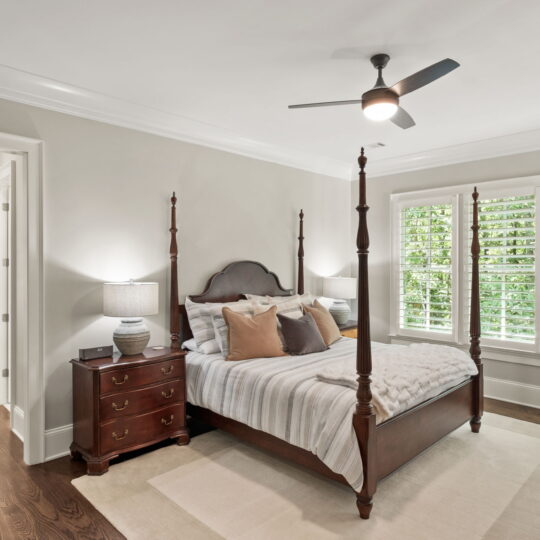 A cozy bedroom with a dark wooden four-poster bed, decorated with pillows and a striped blanket. Two matching nightstands with lamps flank the bed. A ceiling fan hangs above, and large windows with shutters allow natural light to fill the room.