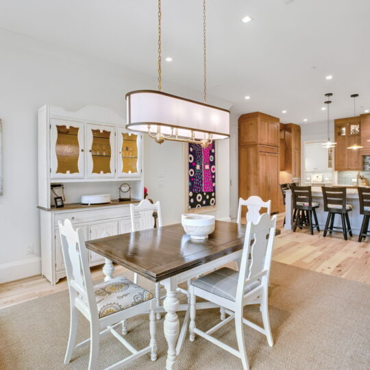 Bright, open dining area with a wooden table and white chairs on a light rug. A large chandelier hangs above. A white cabinet with display shelves sits in the corner. Background shows a kitchen with bar stools and pendant lighting. Light wood flooring.