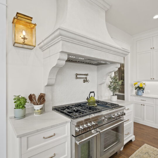 A bright kitchen featuring a large stainless steel stove with a white range hood. There are plants and a kettle on the stove, white cabinets, a wall-mounted lantern, and wooden floors. A hallway is visible to the left.