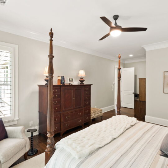 Elegant bedroom with a four-poster bed, striped bedding, and a fur throw. A plush armchair with a purple pillow sits by a shuttered window. A tall wooden dresser holds lamps and decor, and a ceiling fan hangs overhead. A floral painting adorns the wall.