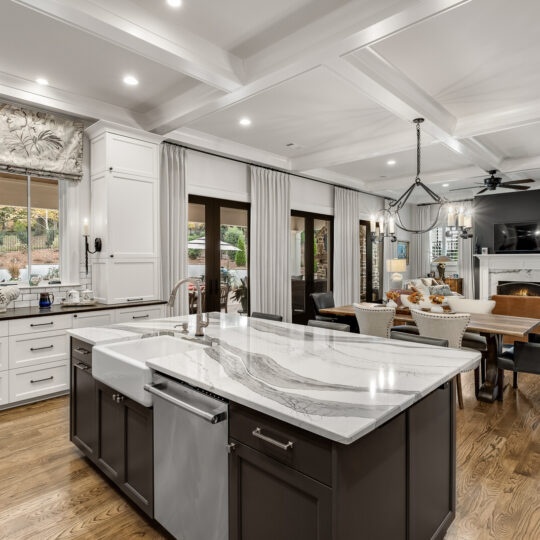 Spacious open-concept kitchen and living area with a large marble island and farmhouse sink in the foreground. Bright hardwood floors, white cabinetry, and ample natural light from large windows and glass doors. Cozy living space with a fireplace and dining area.