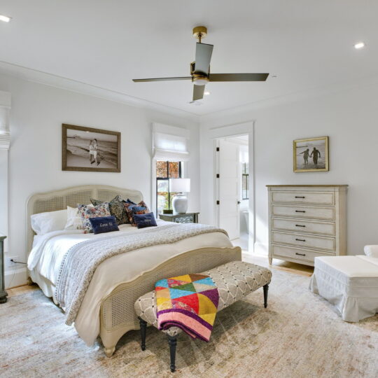 A cozy bedroom with a bed featuring patterned pillows and a colorful quilt at the foot. The room has light walls, a ceiling fan, two windows with blinds, a dresser, an armchair, and decorative artwork on the walls.