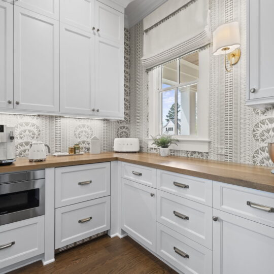 A modern kitchen corner with white cabinets, a wooden countertop, and stainless steel appliances. A coffee machine, mixer, and plant are on the counter. A window with blinds shows a view outside. Patterned wallpaper adds a decorative touch.