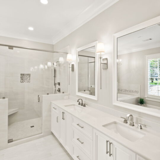 Modern bathroom with dual sinks, large mirrors, and a glass-enclosed shower. White cabinetry, light countertops, and walls create a bright space. Sconce lighting and a window with shutters add elegance.