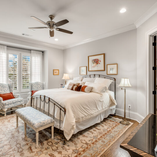 A cozy bedroom with a white bed, patterned rug, and ceiling fan. Two armchairs and a small bench are near a large window with curtains. Wall art decorates the grey walls, and a door leads to another room. A dresser with a mirror is on the right.