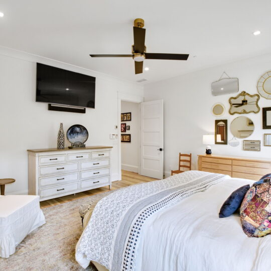 A cozy bedroom featuring a bed with decorative pillows, a white armchair, and a dresser with a TV mounted above. The wall displays various mirrors and art. A ceiling fan hangs from the white ceiling, and soft lighting creates a warm atmosphere.