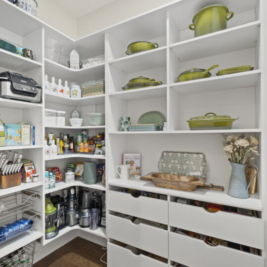 A neatly arranged pantry with white shelves filled with green cookware, various kitchen appliances, and food items. The shelves also hold decorative items and storage baskets, creating an organized and tidy appearance. Wooden floor visible.