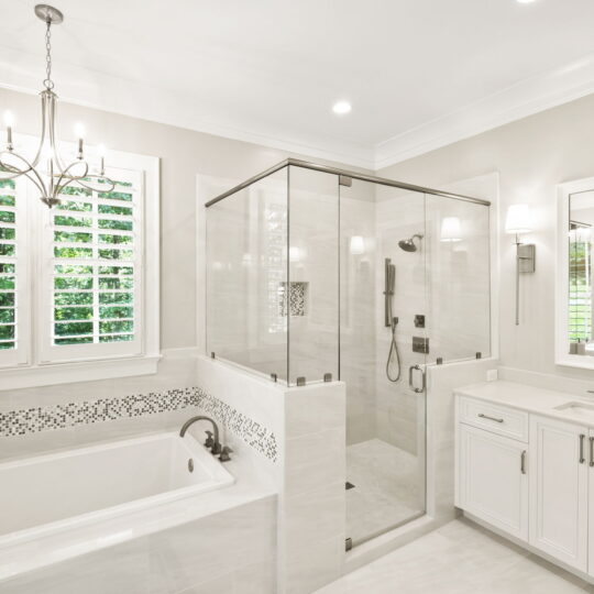 Bright, modern bathroom with white cabinets and walls. Features a large bathtub, glass-enclosed shower, and a double-sink vanity. Natural light enters through a window with shutters. A chandelier hangs from the ceiling, adding elegance.