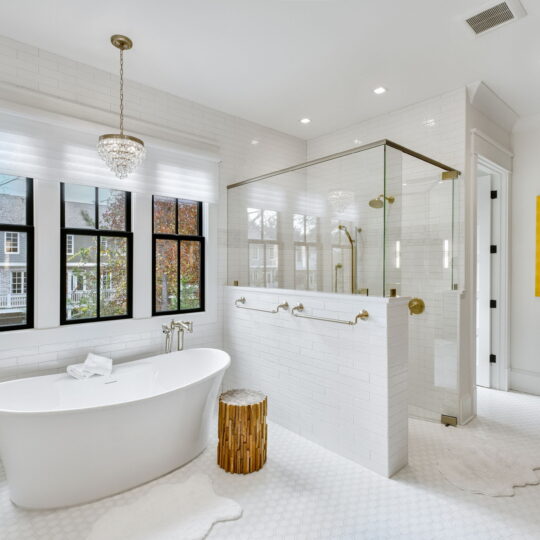 Bright bathroom featuring a freestanding white tub with a wooden stool beside it, a glass-enclosed shower, and black-framed windows. A chandelier hangs above, and a colorful framed artwork is on the wall near the shower. White walls and floor.