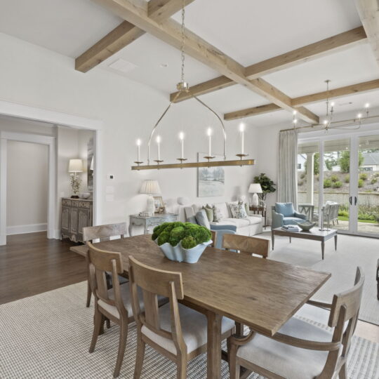 Spacious living and dining area with a wooden beamed ceiling. A wooden dining table with chairs is centered, overlooking a cozy seating area with armchairs. Large windows provide natural light and a view of a landscaped outdoor space.