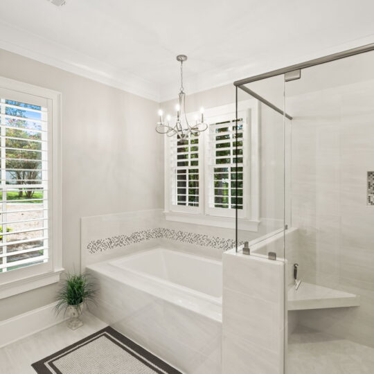 Modern bathroom with a large glass-enclosed shower and a bathtub featuring mosaic tiles. A window with white shutters allows natural light, and a small potted plant decorates the floor. A chandelier hangs from the ceiling, and a rug is near the tub.