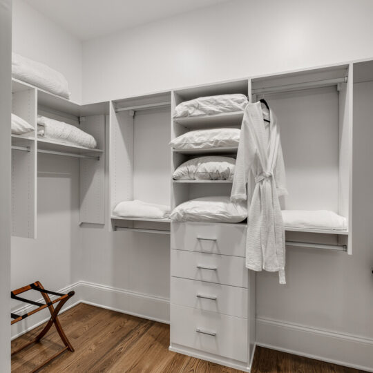 A neatly organized closet with white shelves and drawers. It holds folded linens, a hanging white bathrobe, and an open luggage stand on a wooden floor. The space is well-lit with a modern, minimalist design.