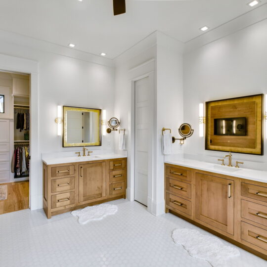 A spacious bathroom features two wooden vanities with sinks, large mirrors, and modern lighting. The white walls and hexagonal tile floor contrast with the warm wood. A walk-in closet is visible through an open door. Two white rugs lie on the floor.