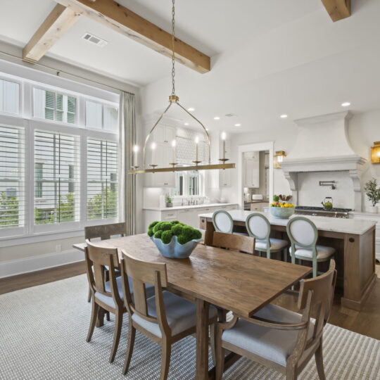 A modern dining room with a wooden table and chairs, a stylish chandelier overhead, and a large window with curtains. Adjacent is a spacious kitchen featuring a white island, bar stools, and a range hood. Light wood floors and exposed beams adorn the ceiling.