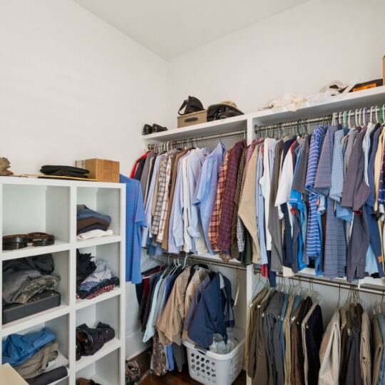 A neatly organized walk-in closet with wooden floors. Clothing is hung on racks, and folded clothes and shoes are stored on shelves. A laundry basket is placed on the floor. The space is well-lit by ceiling light.