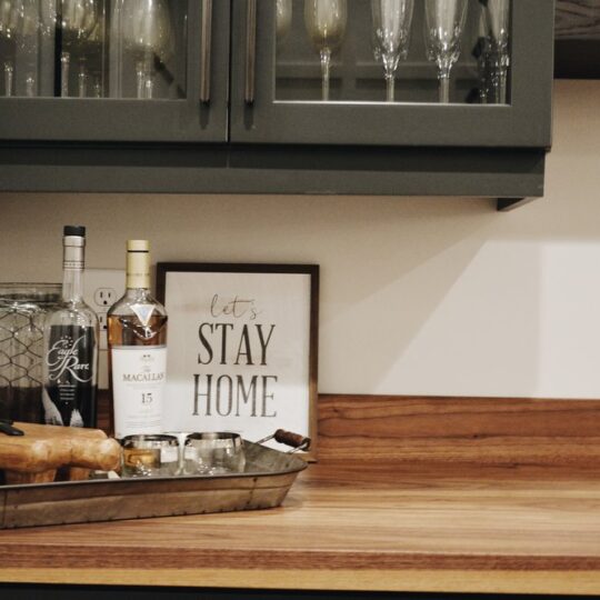 A kitchen counter with a tray holding various bottles and a jar. A framed sign reads Lets Stay Home. Above, glassware is displayed in a cabinet with glass doors. The counter and cabinets have a warm, rustic feel.