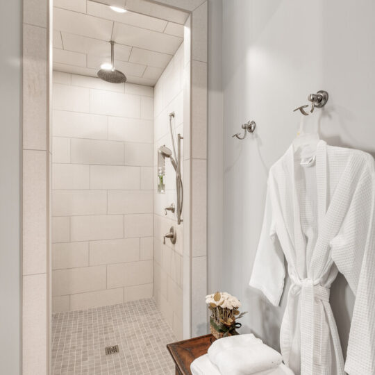 A modern bathroom with a walk-in shower featuring light gray tiles and a rain showerhead. A wooden table holds folded white towels and flowers. A white bathrobe hangs on a wall hook beside the table, and the floor has patterned tiles.